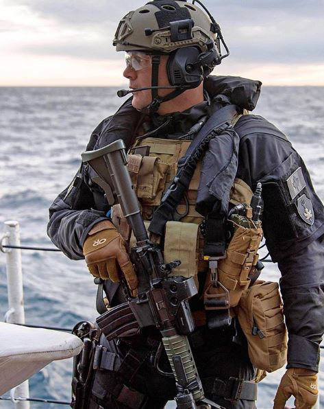 Soldier in tactical gear with a rifle against a sea backdrop.