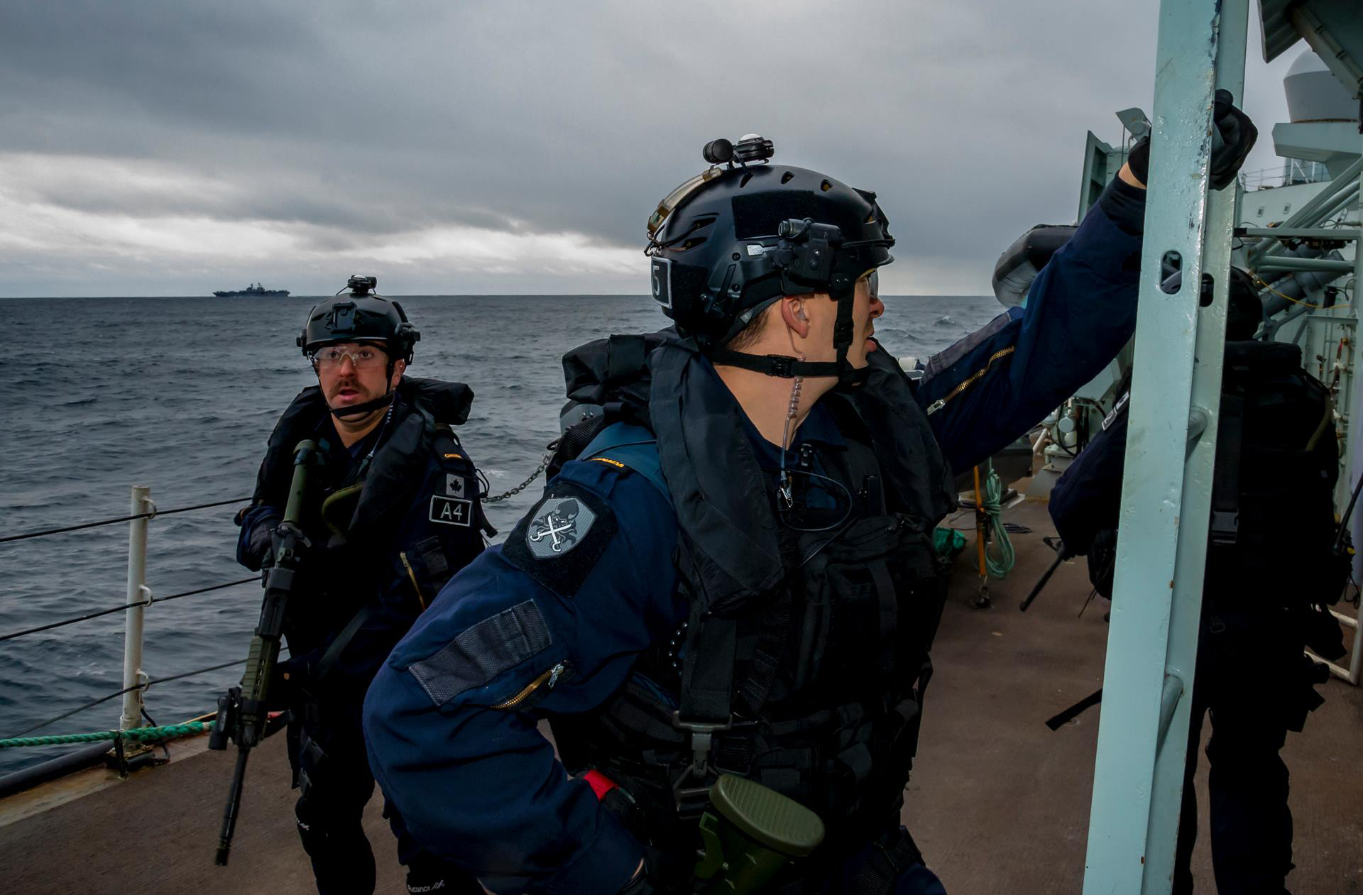 Navy personnel in gear on ship deck with a ship in the distance on overcast sea.