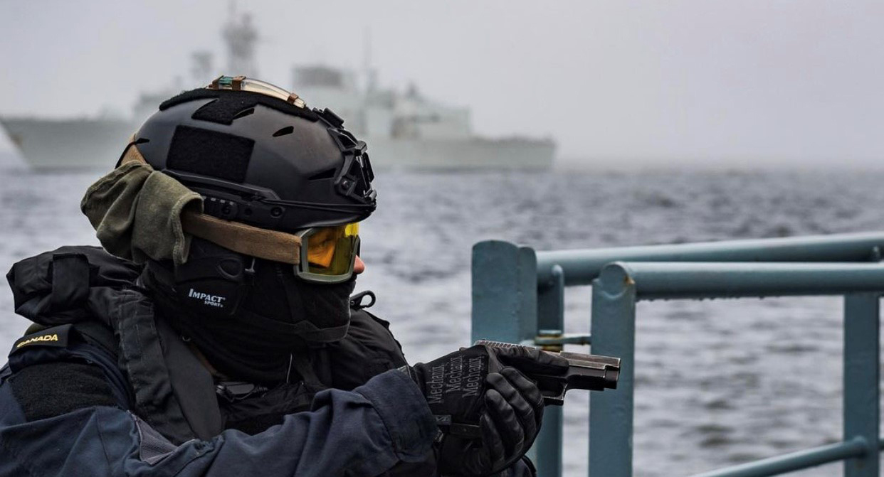 A person in tactical gear holding a handgun on a boat with a ship in the background.