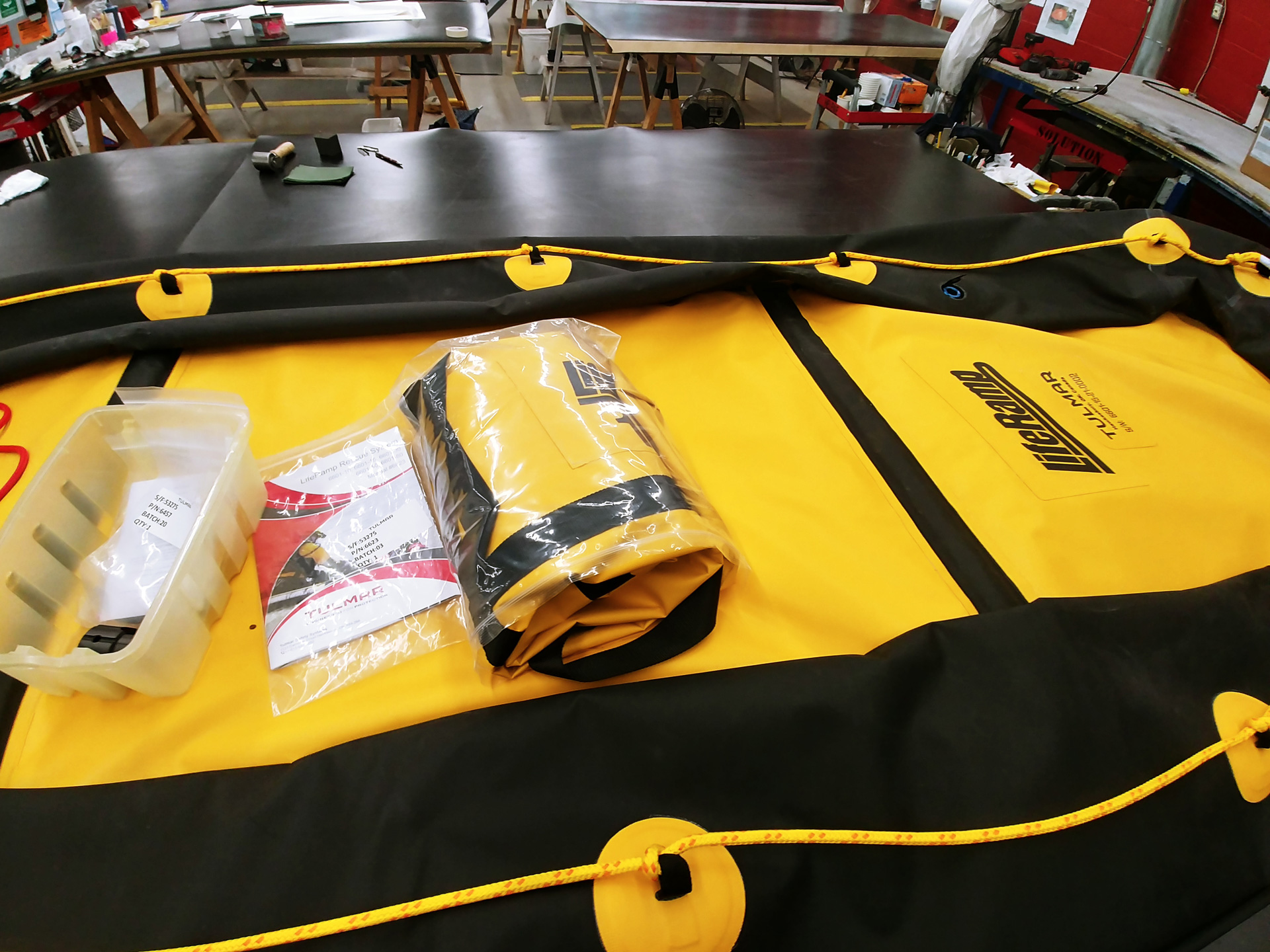 Emergency gear and documents laid on a large yellow and black raft in a manufacturing workshop.