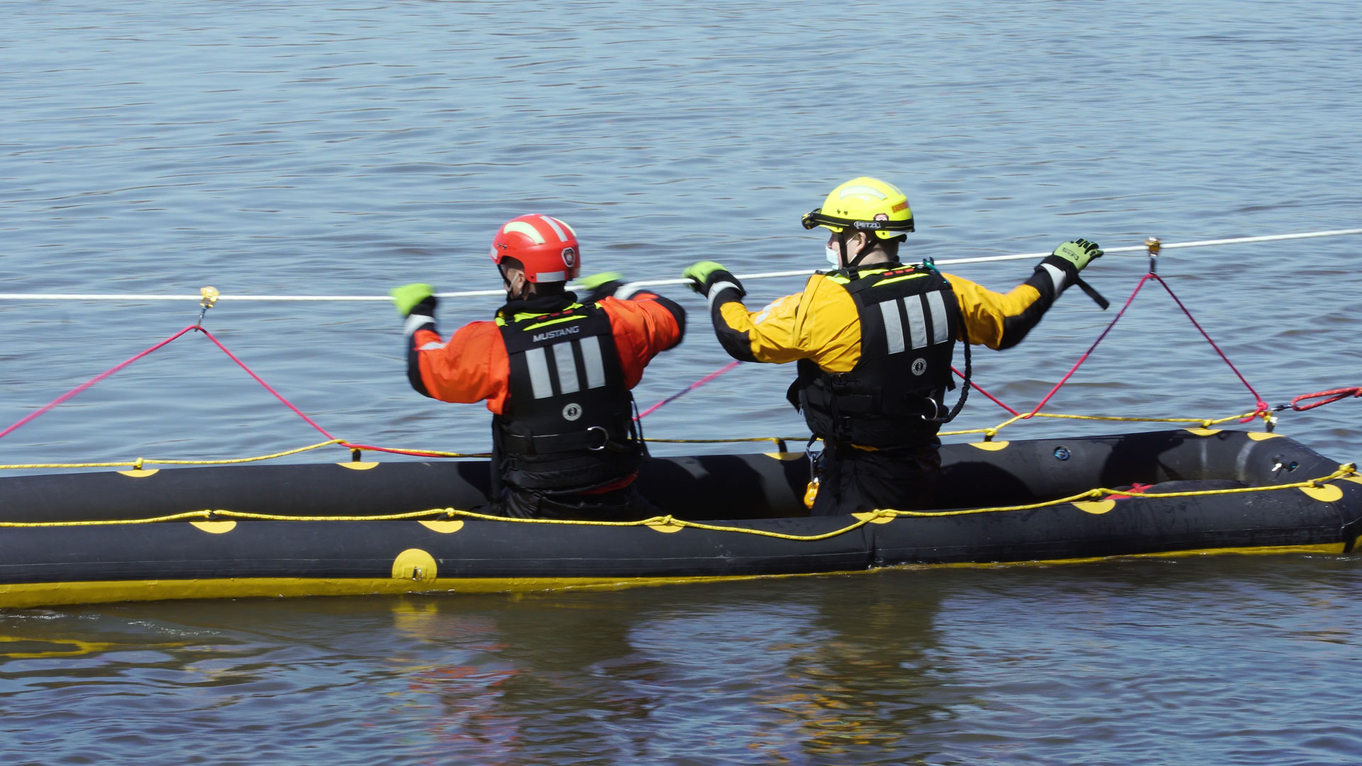 Two rescuers in a raft on the water, holding ropes, wearing safety gear.