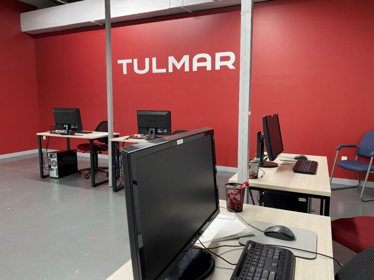 Office space with desks, computers, and a red wall with "TULMAR" written on it.