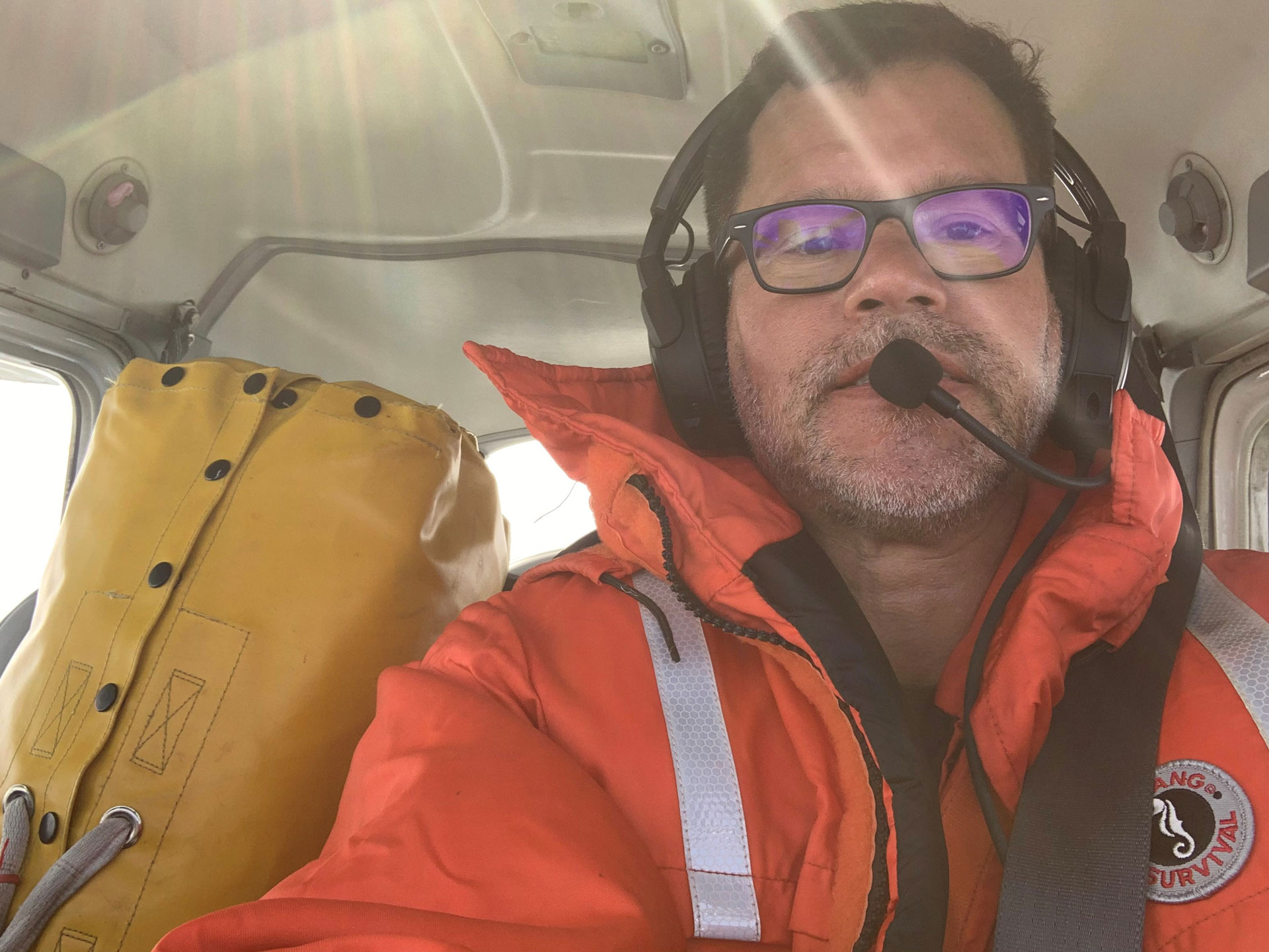 Person in orange survival suit with headset inside vehicle, yellow bag with life raft inside beside them.