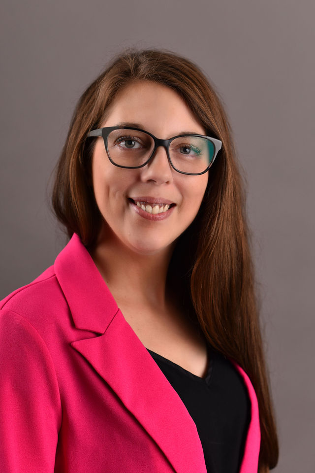 Une femme aux cheveux longs portant un blazer rose et un haut noir, sur fond gris.