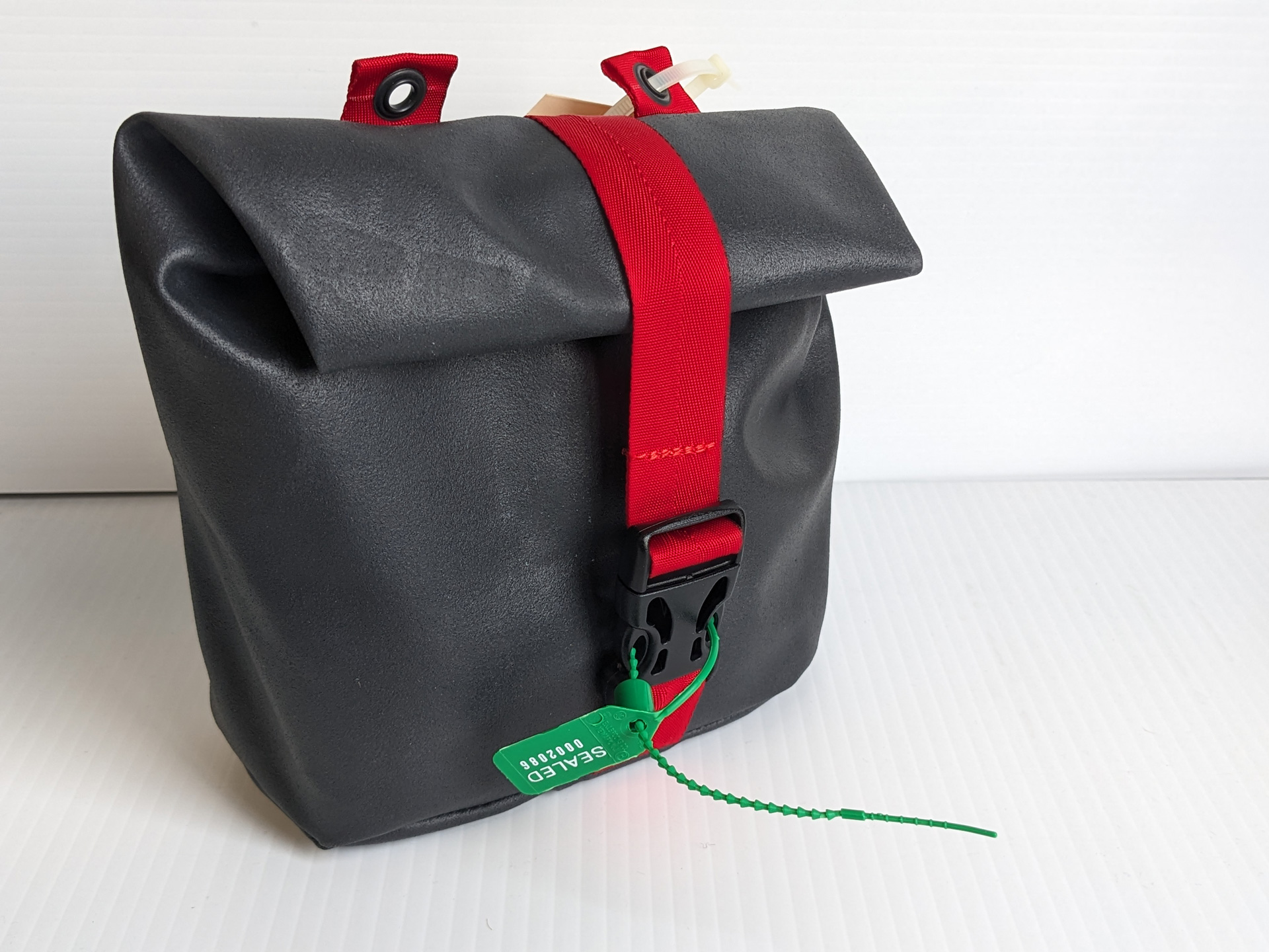 Black roll-top bag with a red strap and a green security tag indicating the tamper-free status of a Passenger Restraint Kit, closed and standing upright.