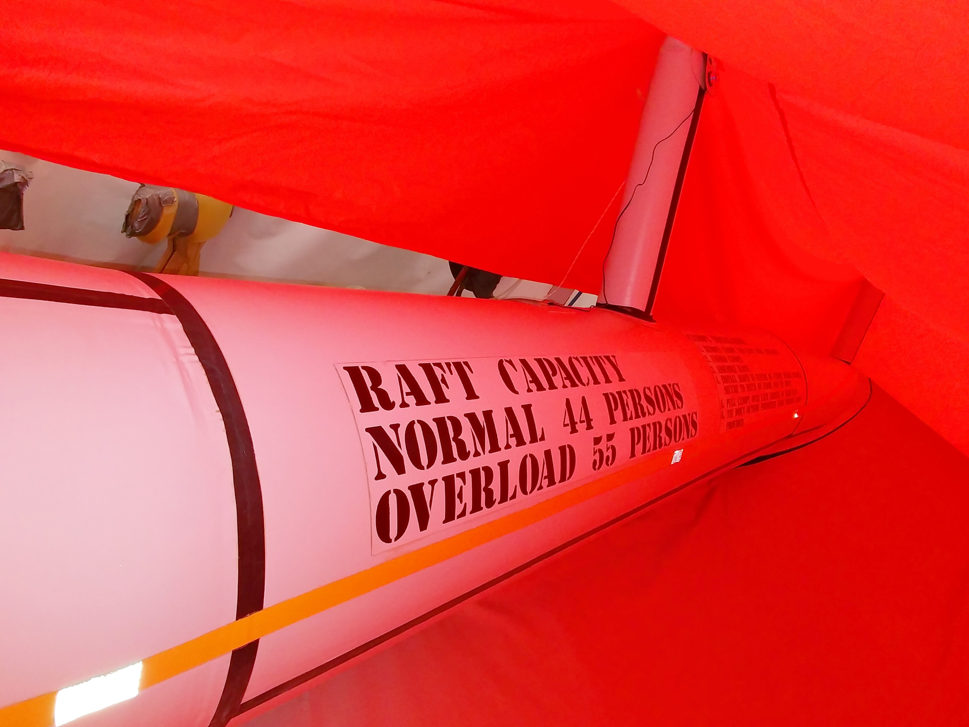 Life raft interior showing capacity label: "NORMAL 44 PERSONS OVERLOAD 55 PERSONS" under red canopy.