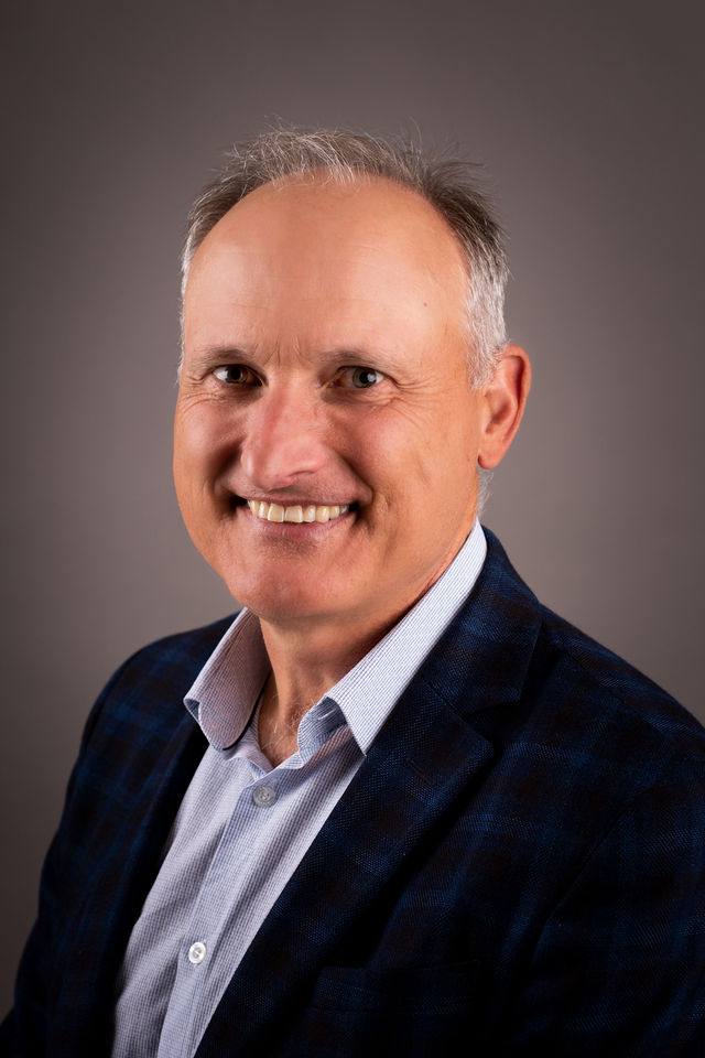 Smiling middle-aged man, Trevor Hains, in a checked navy blazer and light blue shirt, set against a plain grey background, conveying warmth and professionalism.