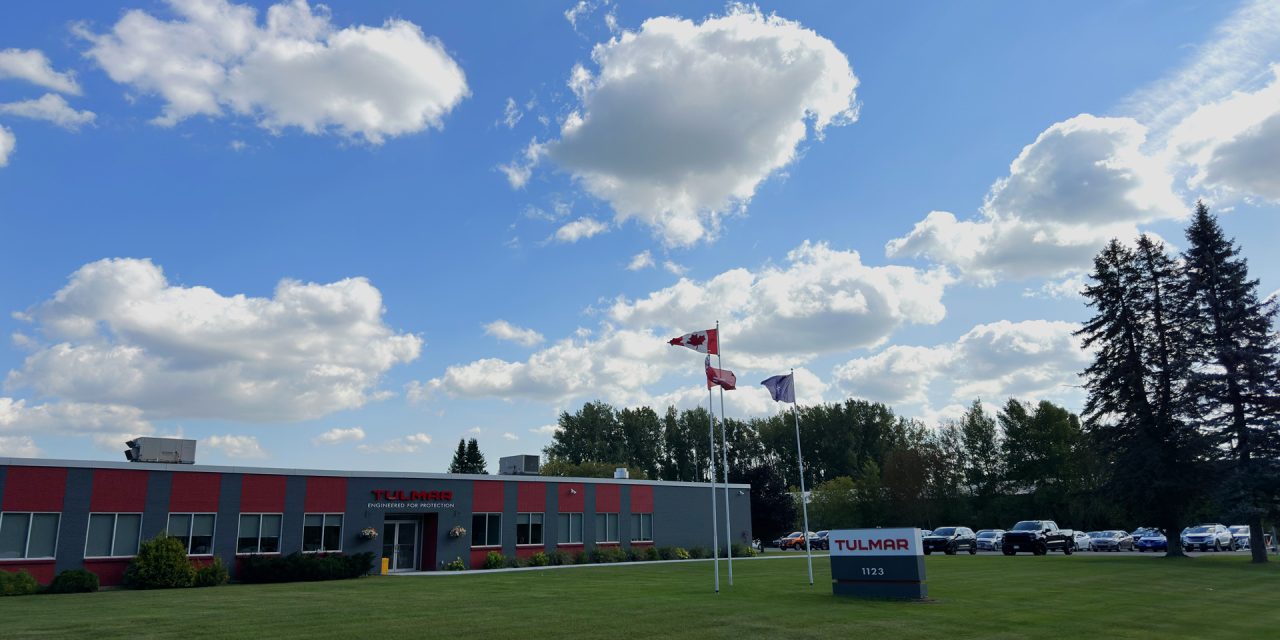 Bâtiment industriel portant le logo "Tulmar", entouré d'une pelouse verte sous un ciel bleu vif avec des nuages cotonneux. Des drapeaux canadiens sont accrochés aux mâts.