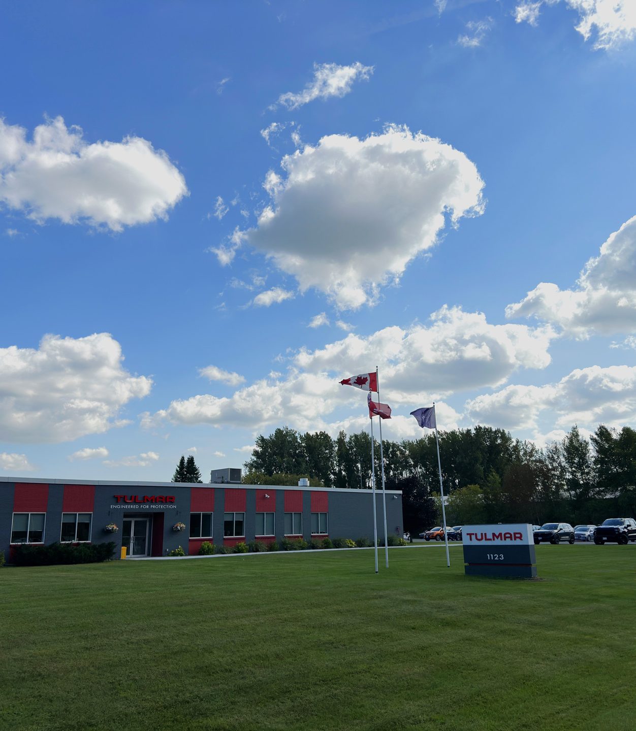 Industrial building with "Tulmar" logo, surrounded by green lawn under a bright blue sky with fluffy clouds. Canadian flags are on flagpoles.